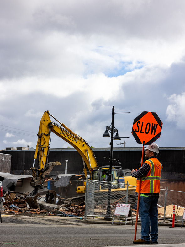 A construction worker holds a "SLOW" sign as a yellow excavator demolishes a building behind a safety fence under a cloudy sky.