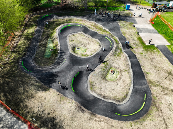 An aerial view shows cyclists riding on a winding paved pump track surrounded by grassy mounds and trees at a park.