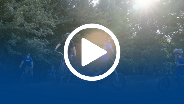 Kids ride bikes through a wooded pump track as sunlight filters through the trees, overlaid with a large play button icon.