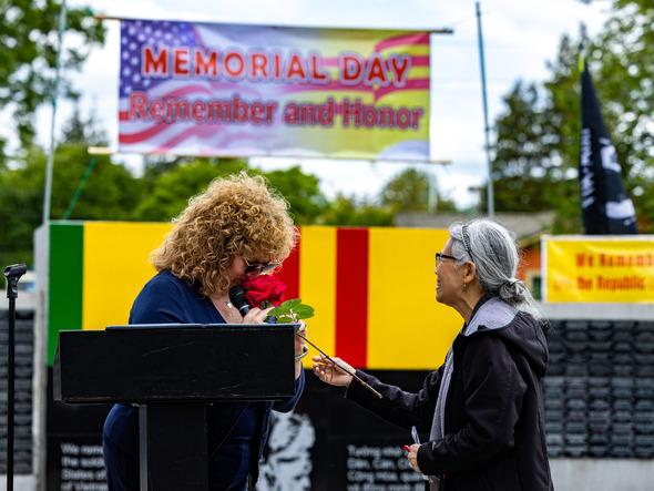 A woman at a podium holds a red rose and bows her head as another woman smiles and offers it during a Memorial Day ceremony.
