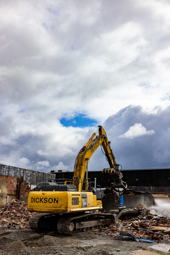 A yellow excavator tears through rubble as demolition continues under a dramatic, cloud-filled sky.