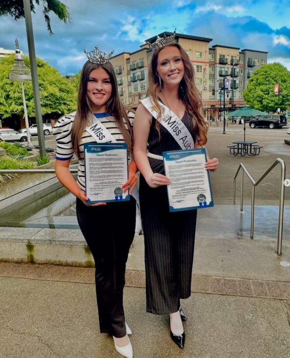 Two titleholders, Miss Auburn and Miss Auburn's Teen, stand outdoors holding official proclamations in front of Auburn's City Hall plaza.