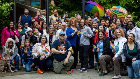 A large, joyful group of people of diverse backgrounds gather outdoors holding Pride flags and umbrellas during a Pride Month celebration.