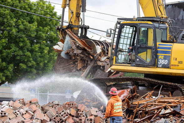An excavator lifts debris while a construction worker sprays water to control dust during a downtown Auburn demolition project.