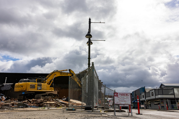 A construction zone in downtown Auburn shows a demolition site with heavy equipment, fencing, and a “No Parking” sign under a dramatic cloudy sky.