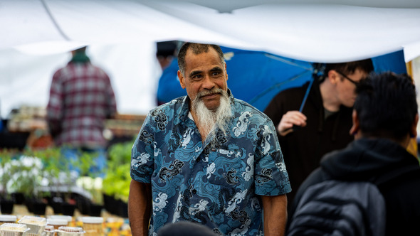A smiling vendor in a blue patterned shirt chats with visitors at a rainy farmers market, surrounded by plants and handmade goods.