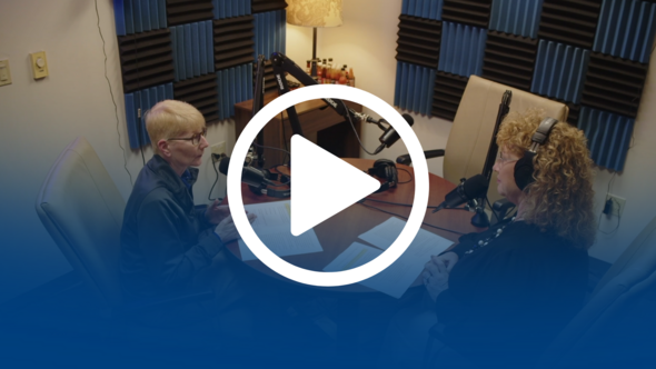 Two people wearing headphones sit across from each other in a podcast studio with microphones, acoustic foam, and papers on the table.