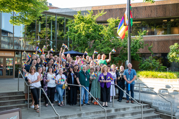 A large group of people cheer and wave Pride flags on the steps outside Auburn City Hall during a Pride flag-raising celebration.