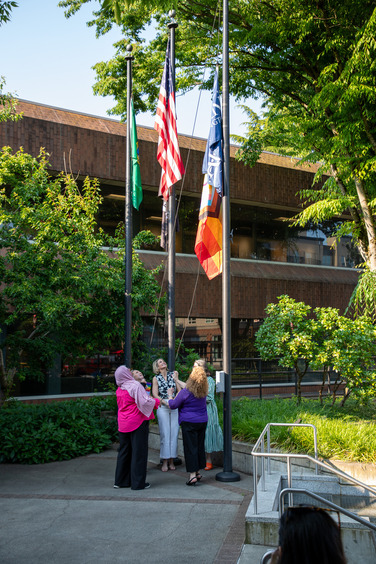 Four people raise a Progress Pride flag outside a government building, standing beneath American and Washington State flags.