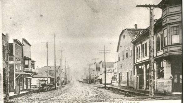 Historic black-and-white photo of a muddy street in early Auburn, WA, lined with wooden buildings, horse-drawn wagons, and tall utility poles.