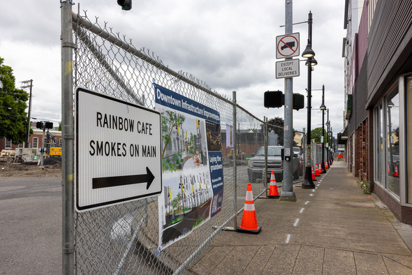 Sidewalk view next to fenced construction zone in downtown Auburn, with signs pointing to Rainbow Cafe and Smokes on Main.