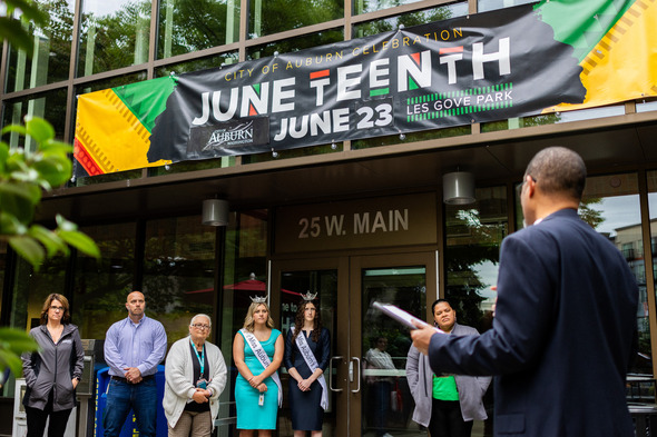 A speaker addresses a small crowd outside Auburn City Hall under a Juneteenth celebration banner.