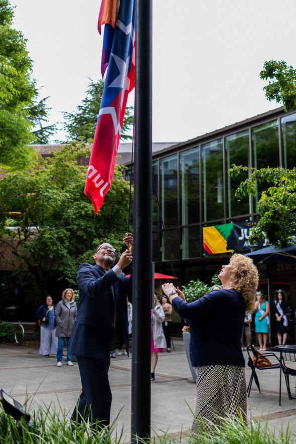 Two people smile as they raise the Juneteenth flag outside Auburn City Hall, with onlookers gathered behind them.