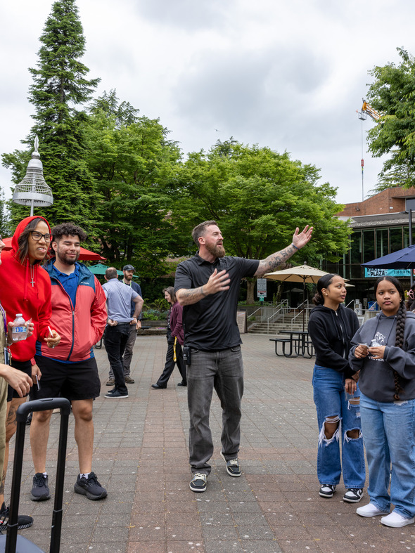 A man gestures while talking to a group of students and staff in a plaza surrounded by trees.