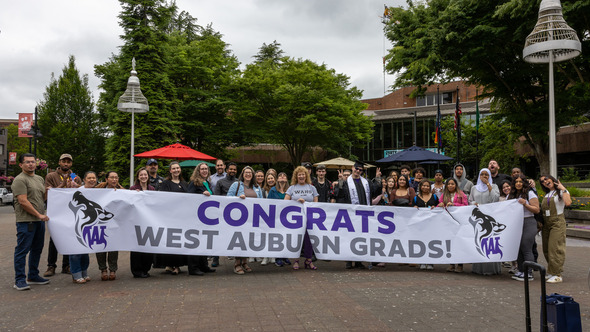 A large group poses behind a “Congrats West Auburn Grads!” banner in front of Auburn City Hall.