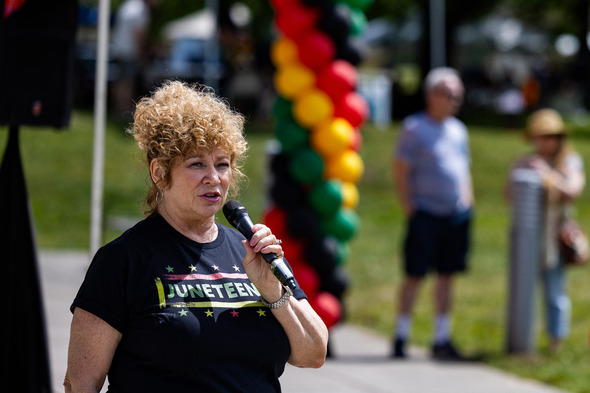 Woman speaks into a microphone at a Juneteenth event, standing in front of red, yellow, green, and black balloon columns.
