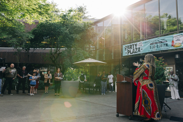 A speaker in a vibrant red patterned dress addresses a crowd at City Hall Plaza during a Juneteenth celebration.