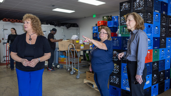 Three women talking inside a food storage room filled with colorful crates and supplies.