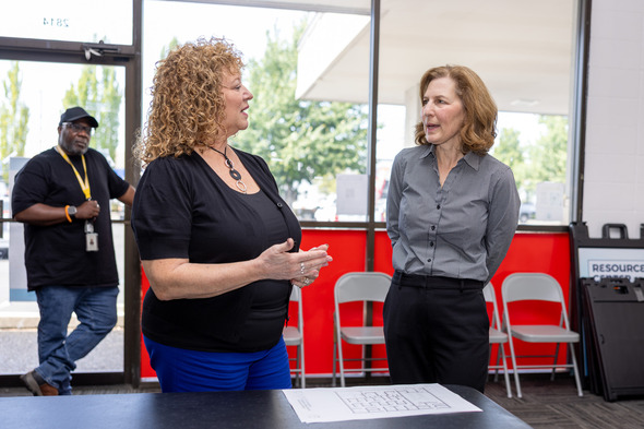 Two women speaking near a table with a layout plan, a man stands in the background near the door.