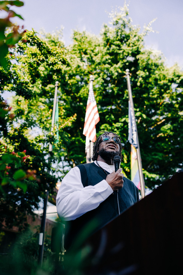 A man speaks into a microphone at a podium outdoors, with flags and trees in the background.