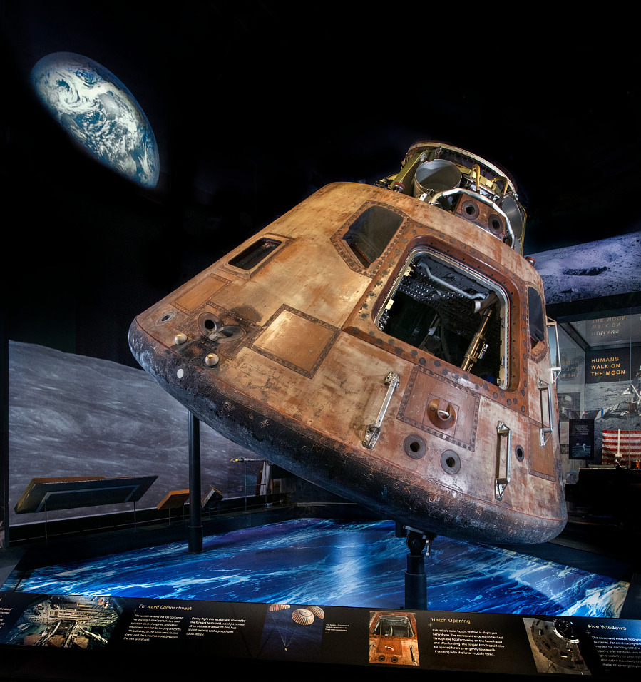 Apollo 11 Command Module on display with Earth and Moon visuals in background, showcasing its historic return from the first Moon landing.