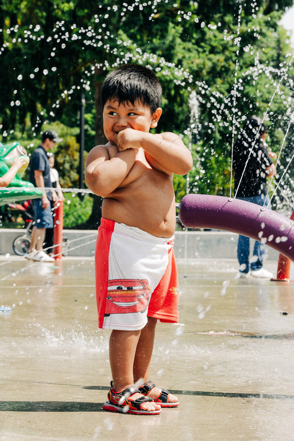 Young boy in red Lightning McQueen shorts playing in a splash pad, smiling as water sprays around him.
