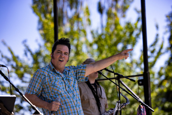 A singer onstage in a plaid shirt points into the crowd while performing during a sunny outdoor concert.