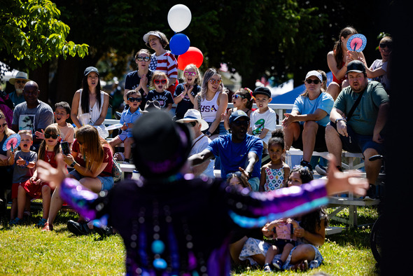 Performer entertains a crowd of kids and families seated on bleachers at an outdoor Fourth of July event.