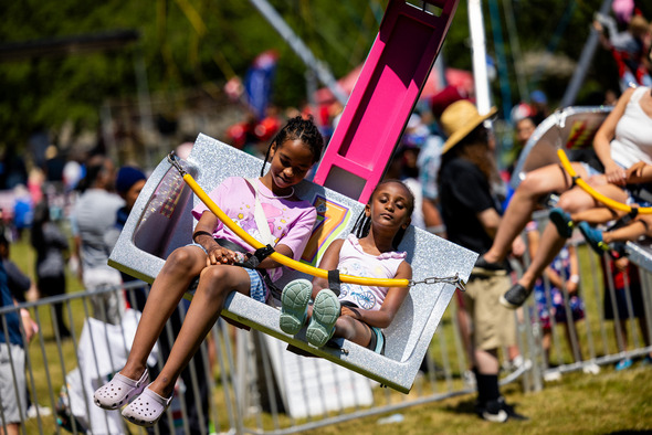 Two young girls ride a carnival swing ride, smiling and relaxed, surrounded by a crowd enjoying the fair.