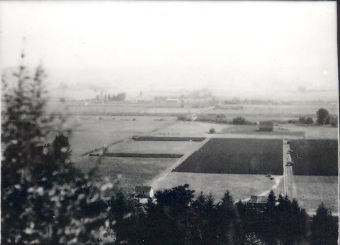 Black and white aerial photo of rural farmland with open fields, dirt roads, and a few scattered buildings surrounded by trees.