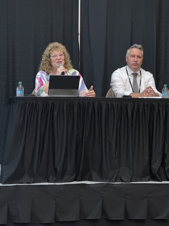 Two speakers at a panel discussion; the woman speaks while the man listens attentively.