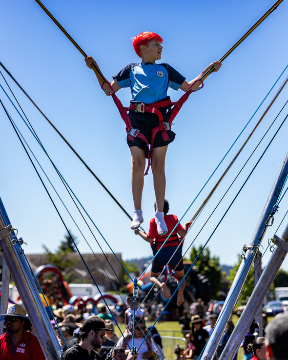 A boy with red hair bounces high on a harnessed trampoline at a summer festival.