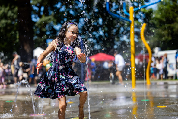 A young girl joyfully plays in a splash pad with water spraying around her at a park.