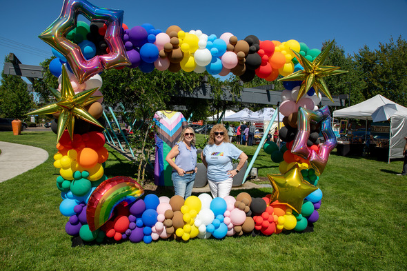 Two women stand smiling inside a balloon arch decorated with rainbow stars and shapes at a Pride event.