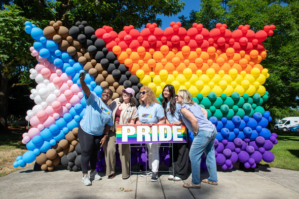 A group poses for a selfie in front of a large balloon wall featuring the Pride flag colors.