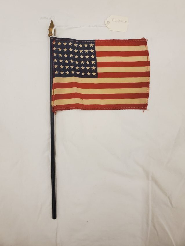 Small vintage American flag with 48 stars, mounted on a black stick with a gold spear tip and white tag attached.