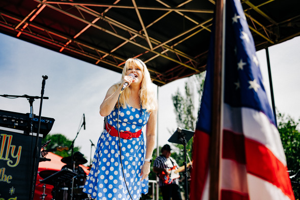 Singer in blue polka-dot dress performing on outdoor stage next to American flag.