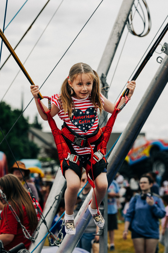 Young girl in patriotic outfit bouncing on a bungee trampoline at a festival.