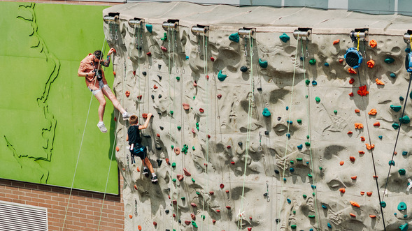 A photo of a man being belayed at a climbing wall outside, taking a photo of a kid climbing up the wall 