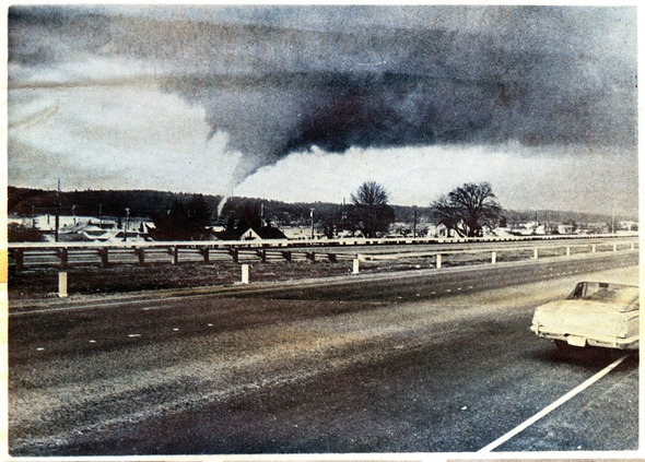 A tornado touches down near residential buildings, viewed from a highway with a vintage car in the foreground.
