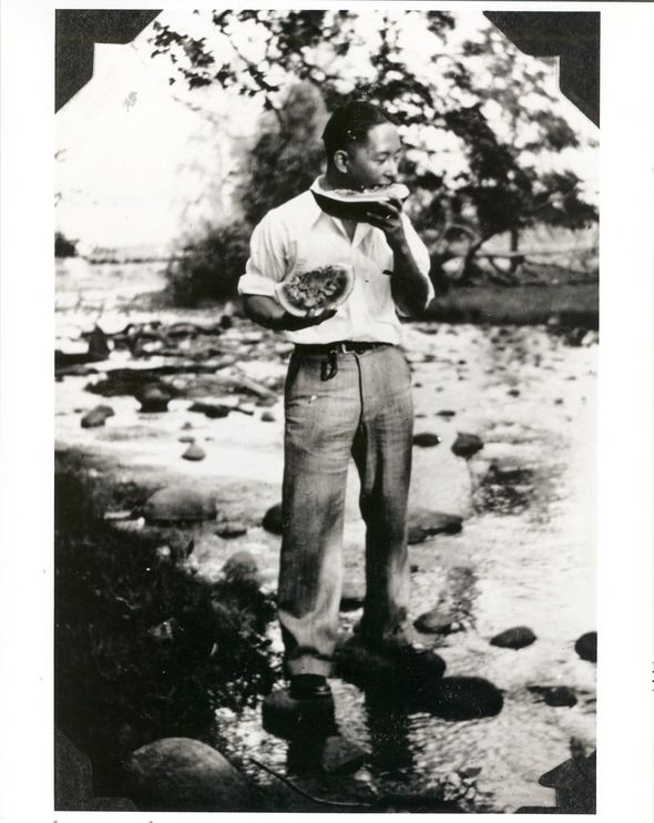 A man stands in a shallow stream eating watermelon, holding one slice and biting into another, with trees and rocks in the background.