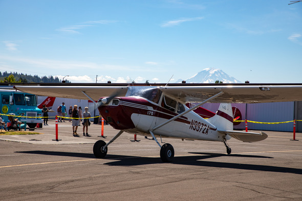 A small red and white plane is parked on the tarmac at an airport event, with people nearby and Mount Rainier visible in the background.