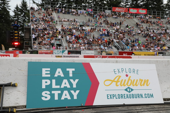 View of packed grandstands behind an Explore Auburn banner at a racetrack.