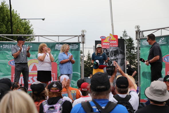 Group on NHRA stage clapping during event, crowd watching from below.