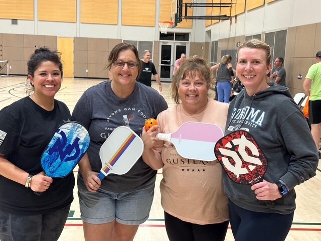 Four women smiling in a gym holding colorful pickleball paddles.