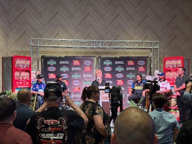 A woman standing at a podium on a stage while drivers surround her at the NHRA media event