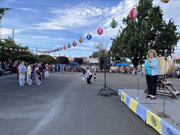 Mayor Backus speaks on stage at the Bon Odori Festival, with dancers and audience in background.