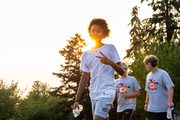 Teens walk a 5K route at sunset, one flashing a peace sign and holding water, with “Almost 5K Poker Run” shirts in the background.