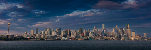 Panoramic view of the Seattle skyline at dusk, featuring the Space Needle and downtown buildings under a dramatic cloudy sky.