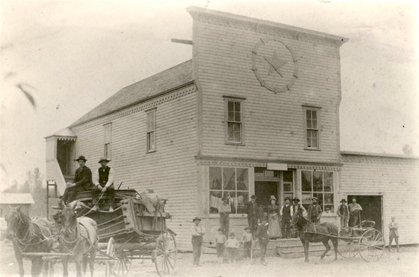 Historic black-and-white photo of early Auburn residents outside a wooden building with a star emblem, horse-drawn wagons parked out front.